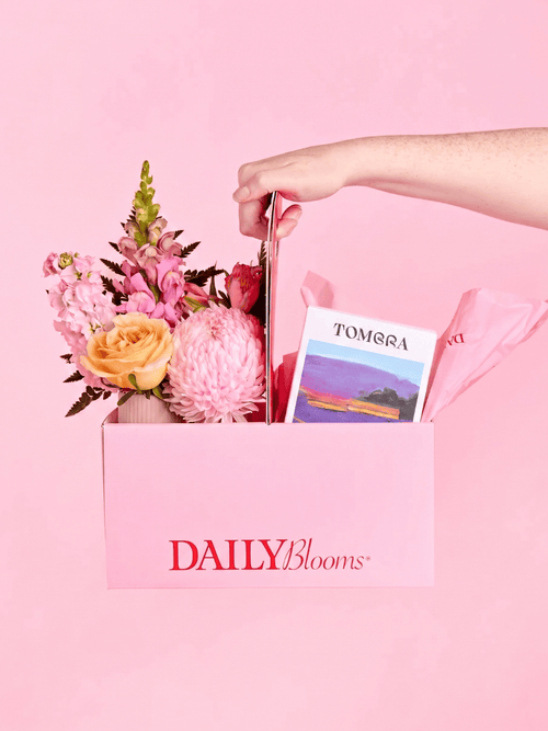 A hand holds a pink DailyBlooms gift box filled with pink and peach flowers, a small card, and pink tissue paper against a solid pink background.