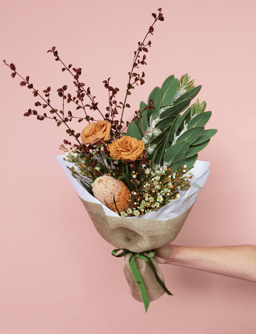 A  loral bouquet held by a person's hand against a solid dusty pink background. The bouquet features orange roses, green leaves, small white baby's breath, and dark reddish-brown berry branches, wrapped in white tissue paper and natural hessian with a green ribbon.
