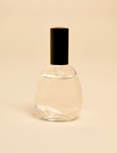 A clear glass room spray bottle with a black cylindrical cap, containing a pale liquid, set against a solid beige background.
