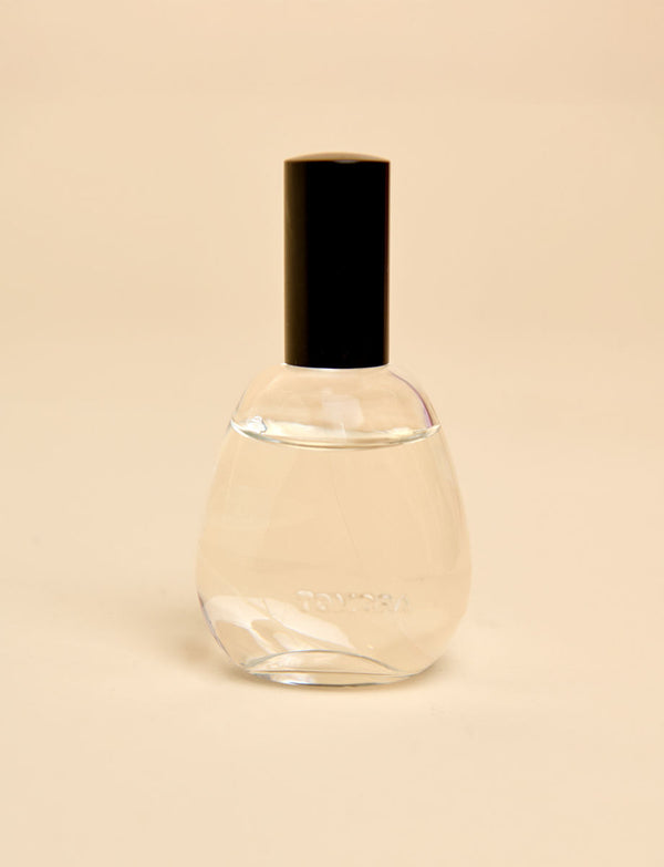A clear glass room spray bottle with a black cylindrical cap, containing a pale liquid, set against a solid beige background.